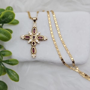 Può includere: Collana con pendente a croce dorata dal design dettagliato. La croce presenta pietre preziose rosse ovali e cristalli trasparenti, incastonati in una montatura dorata. La catena della collana è a maglia piatta e delicata.