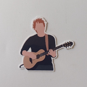 Könnte beinhalten: Ein Aufkleber mit einer Person mit roten Haaren, die eine braune Akustikgitarre spielt.