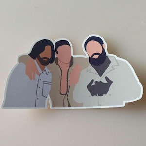 Op de afbeelding: Een sticker met een gestileerde illustratie van drie mensen die elkaar omarmen. De figuren zijn in neutrale tinten, met één persoon die een lichtblauw overhemd draagt, een ander een bruin overhemd en de derde een witte jas.