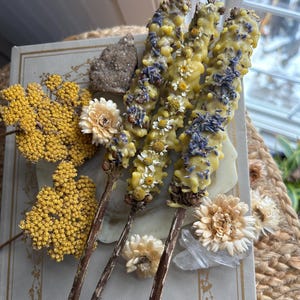Op de afbeelding: Drie houten stokjes bedekt met gele was en ingelegd met gedroogde lavendelknoppen en kleine witte kamillebloemen. De stokjes liggen op een lichtgekleurd boek, samen met trossen gele duizendblad, gedroogde witte bloemen en kleine kristallen.