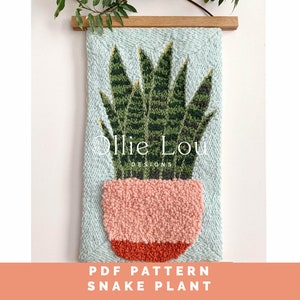 May include: A woven wall hanging with a snake plant design. The plant has green and yellow leaves in a pink pot with an orange base, set against a light blue background. The text "Ollie Lou DESIGNS" and "PDF PATTERN SNAKE PLANT" are visible.