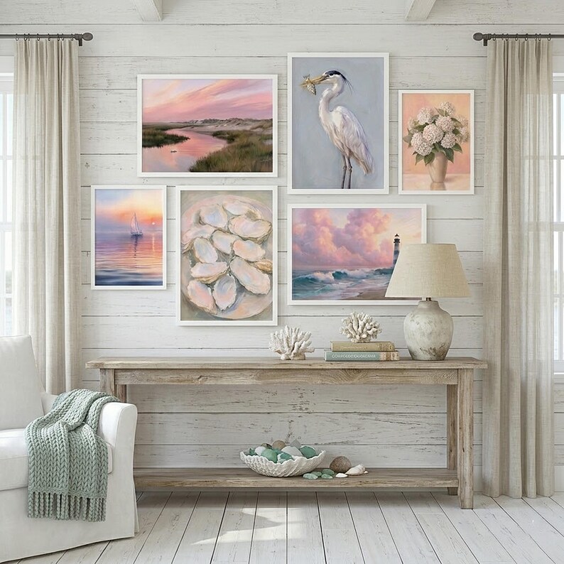 Op de afbeelding: Een galerijwand met kustthema met ingelijste kunstdrukken. De kunstwerken omvatten een zeilboot op het water, een reiger met een vis, een landschap met een vuurtoren en een stilleven van oesters. Een houten console met een lamp en decoratieve koraalaccenten maakt de sc&egrave;ne compleet.