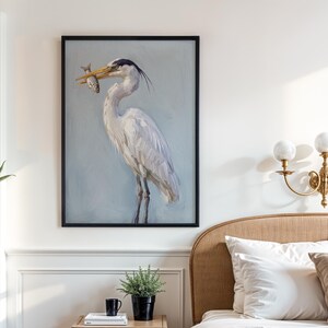 K&ouml;nnte beinhalten: Gerahmtes Gem&auml;lde eines Reihers, der einen Fisch im Schnabel h&auml;lt. Der Reiher ist in Wei&szlig;- und Graut&ouml;nen vor einem hellblauen Hintergrund dargestellt. Das Kunstwerk h&auml;ngt an einer wei&szlig;en Wand &uuml;ber einem Bett mit wei&szlig;en Kissen und einem beigen Kopfteil.