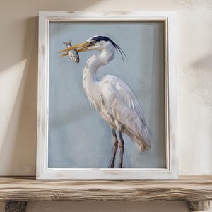 K&ouml;nnte beinhalten: Gerahmtes Gem&auml;lde eines wei&szlig;en Reihers mit einem Fisch im Schnabel. Der Reiher hat einen schwarz-blauen Kopf und ist vor einem hellblauen Hintergrund platziert. Der Rahmen ist wei&szlig; mit einem Used-Look. Das Gem&auml;lde steht auf einem Holzregal.