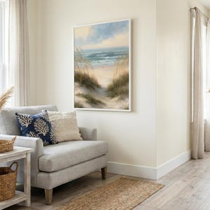 May include: A rectangular wall art print featuring a coastal landscape with sand dunes, tall beach grass, and a calm ocean under a soft blue and yellow sky, framed in a thin white border, displayed on a light-colored wall above a gray armchair.