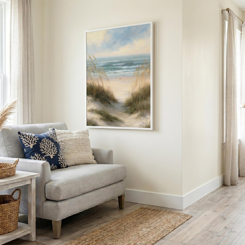 May include: A rectangular wall art print featuring a coastal landscape with sand dunes, tall beach grass, and a calm ocean under a soft blue and yellow sky, framed in a thin white border, displayed on a light-colored wall above a gray armchair.