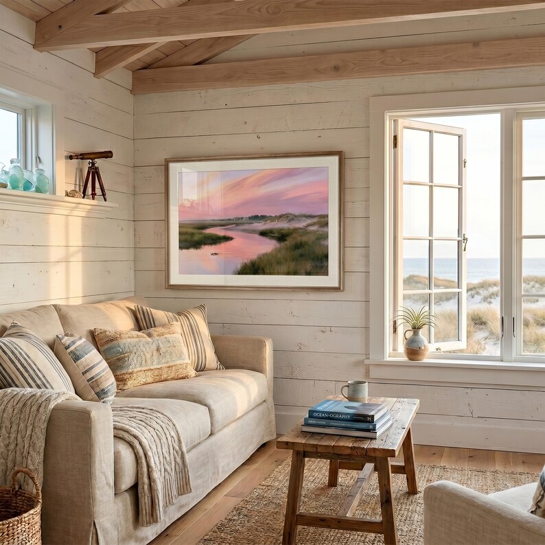 Op de afbeelding: Een gezellige woonkamer met een beige bank, kussens en een deken. Een ingelijst landschapsschilderij hangt boven de bank. Een kleine houten tafel toont boeken en een mok. Een raam biedt uitzicht op de zee en een strand.