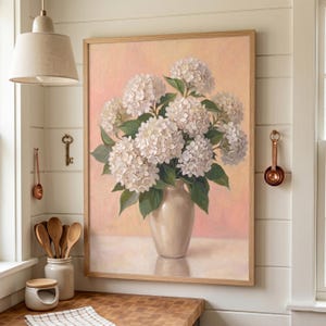 Op de afbeelding: Een ingelijst schilderij van witte hortensia's in een cr&egrave;mekleurige vaas. Het kunstwerk heeft een zachte, schilderachtige stijl met een roze en perzikkleurige achtergrond. De houten lijst completeert het bloemstuk.