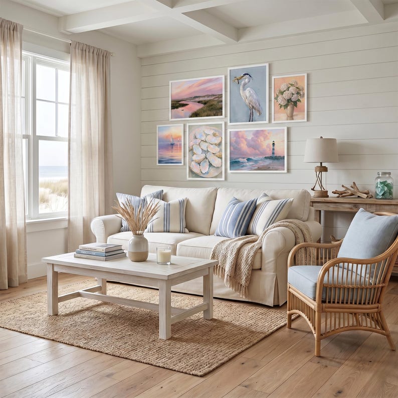 Op de afbeelding: Een woonkamer met een kustthema, met een witte bank, blauw-wit gestreepte kussens en een geweven vloerkleed. Kunstwerken aan de muur omvatten landschapsschilderijen en een schilderij van een reiger. Een witte salontafel en een rotan stoel zijn ook aanwezig.