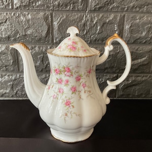 May include: A white porcelain teapot with a floral pattern of pink roses and green leaves. The teapot has a gold trim around the lid and spout.