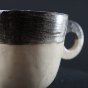 May include: A white ceramic mug with a black glaze around the top. The mug has a crackled glaze and a handle.