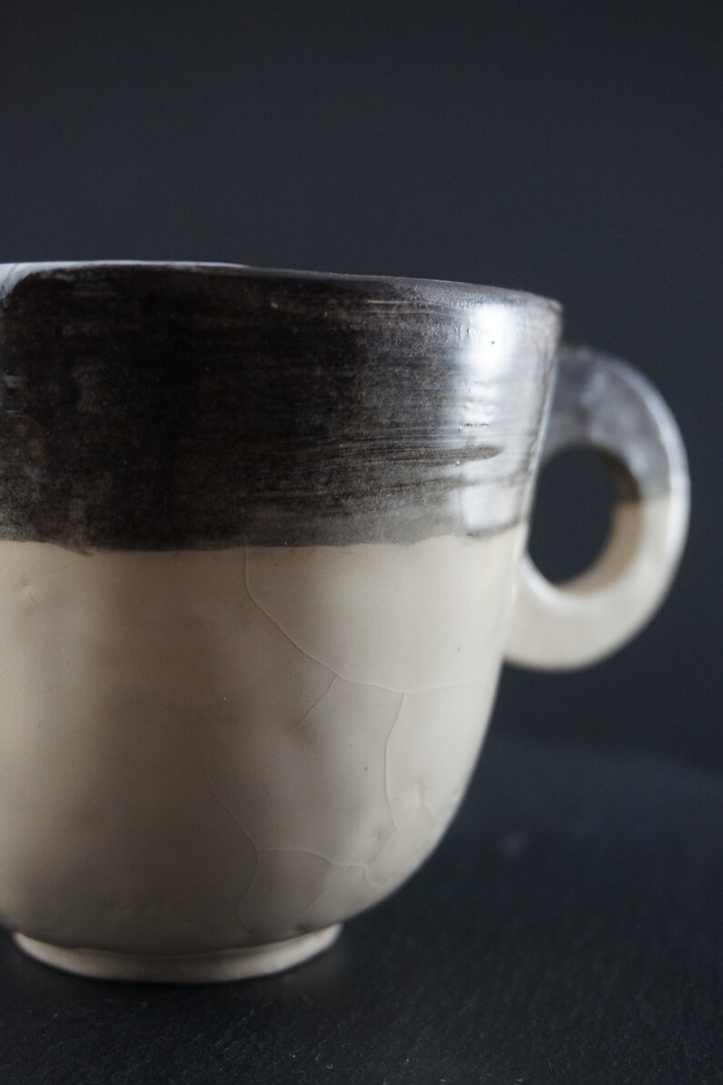 May include: A white ceramic mug with a black glaze around the top. The mug has a crackled glaze and a handle.