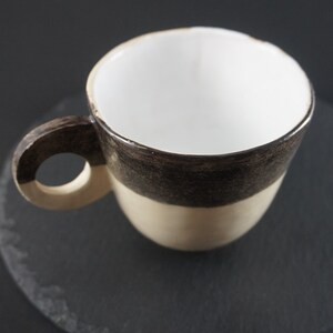 May include: A white ceramic coffee cup with a brown and beige glaze. The cup has a rounded handle and is sitting on a black slate coaster.