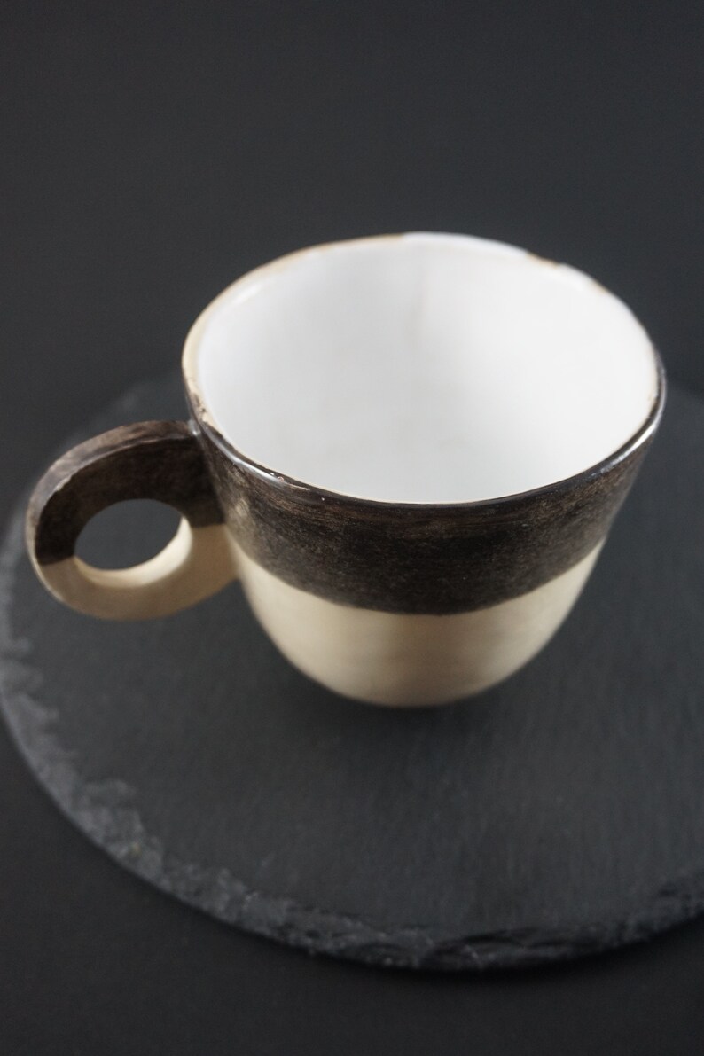 May include: A white ceramic coffee cup with a brown and beige glaze. The cup has a rounded handle and is sitting on a black slate coaster.
