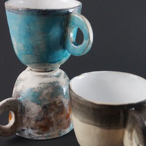 May include: Two ceramic mugs, one stacked on top of the other. The top mug is a light blue color with a white interior and a rounded handle. The bottom mug is a light brown color with a white interior and a rounded handle. The mugs have a rustic, handmade look.