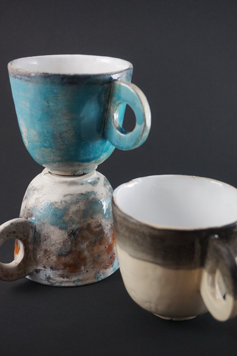 May include: Two ceramic mugs, one stacked on top of the other. The top mug is a light blue color with a white interior and a rounded handle. The bottom mug is a light brown color with a white interior and a rounded handle. The mugs have a rustic, handmade look.