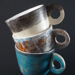 May include: Three ceramic mugs stacked on top of each other. The top mug is white with black and gray stripes. The middle mug is brown with white and orange speckles. The bottom mug is teal blue with a white glaze.