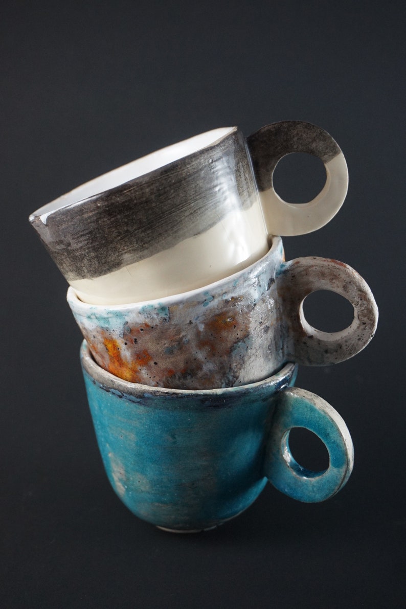 May include: Three ceramic mugs stacked on top of each other. The top mug is white with black and gray stripes. The middle mug is brown with white and orange speckles. The bottom mug is teal blue with a white glaze.