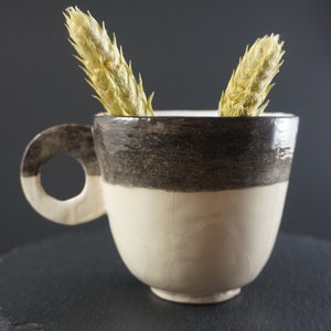 May include: A white ceramic cup with a brown band around the middle. The cup has a handle and is sitting on a black slate coaster. Two dried flower buds are sticking out of the cup.
