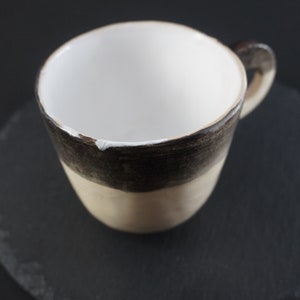 May include: A white ceramic mug with a brown and beige glaze. The mug has a rounded shape and a handle.