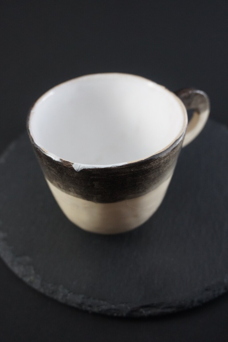 May include: A white ceramic mug with a brown and beige glaze. The mug has a rounded shape and a handle.