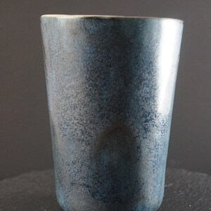 Op de afbeelding: Een blauwe keramische beker met een gespikkeld glazuur. De beker heeft een ronde bodem en een rechte, brede opening.