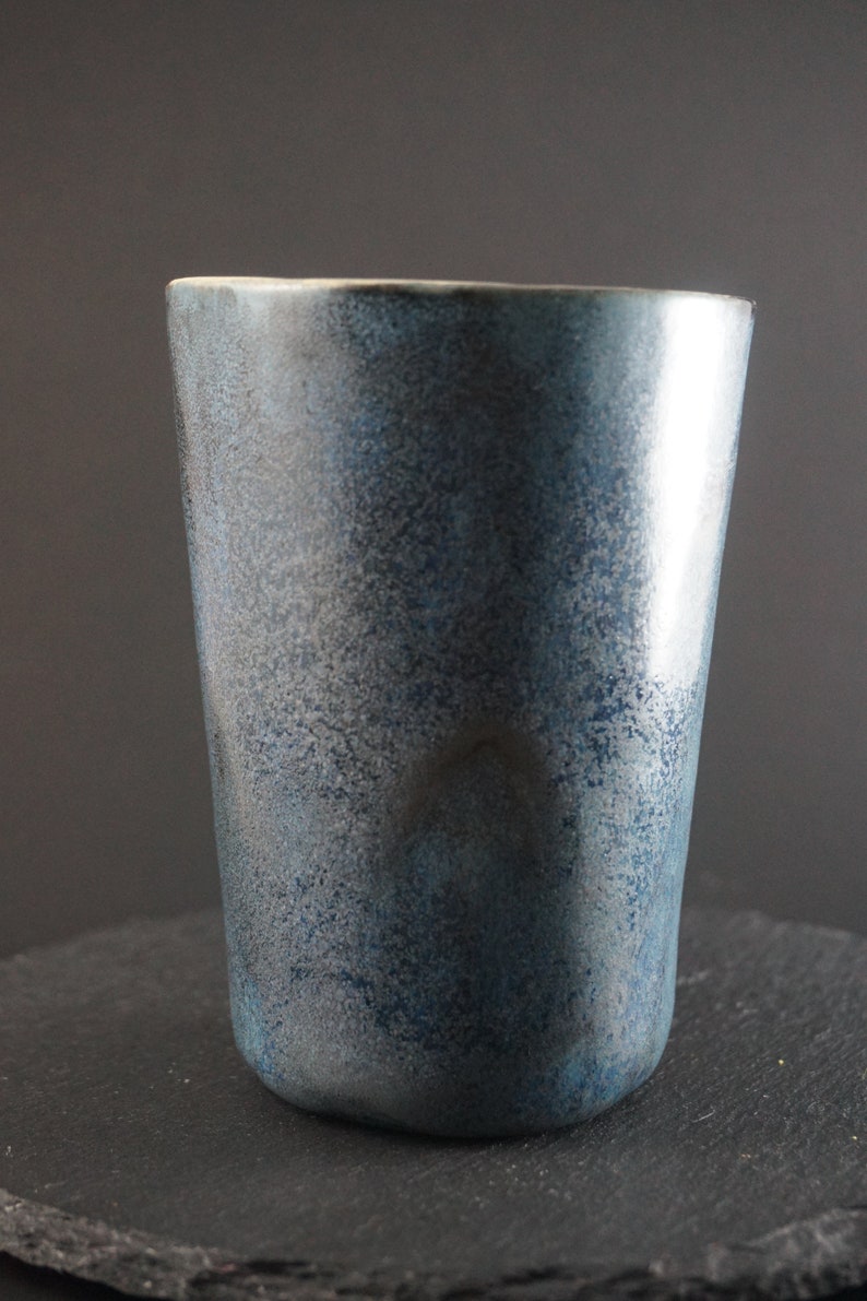 Op de afbeelding: Een blauwe keramische beker met een gespikkeld glazuur. De beker heeft een ronde bodem en een rechte, brede opening.