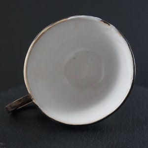 May include: A white ceramic teacup with a black rim and a gold accent. The cup is sitting on a black slate surface.