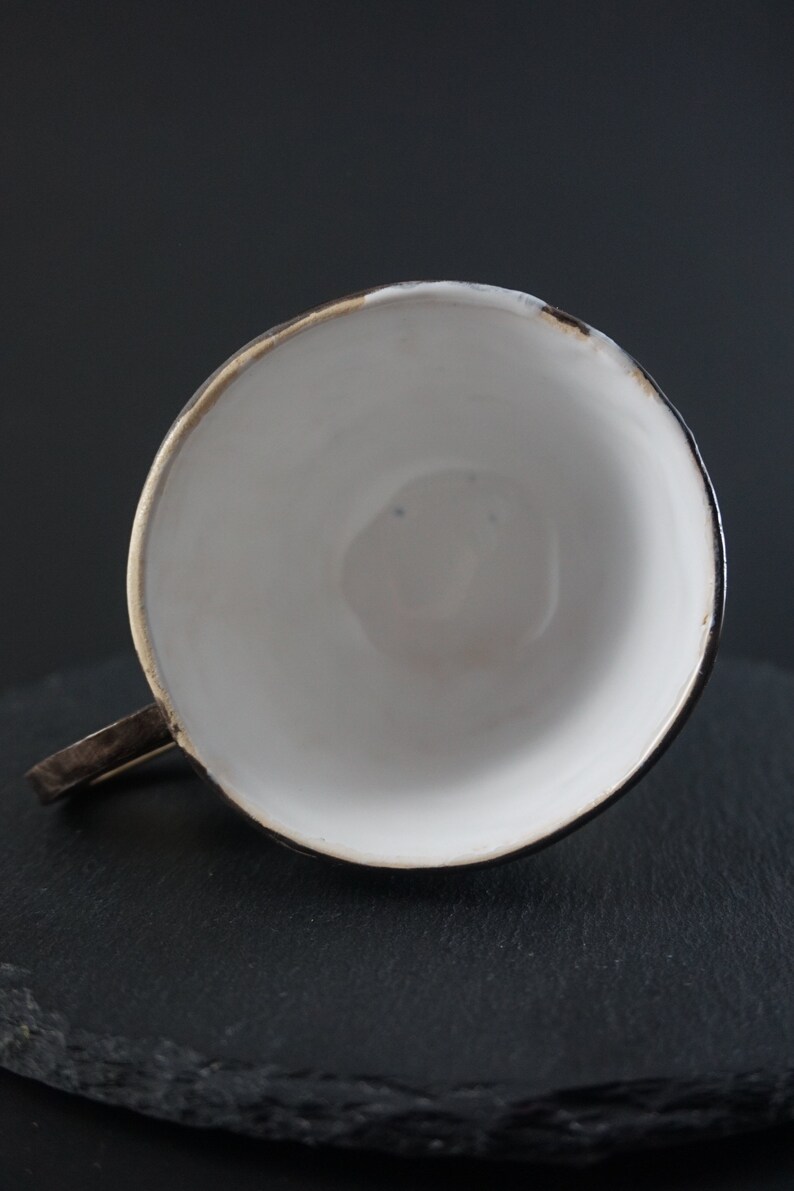 May include: A white ceramic teacup with a black rim and a gold accent. The cup is sitting on a black slate surface.