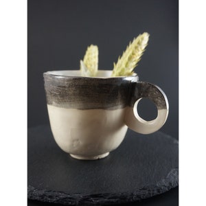 May include: A ceramic mug with a black and white glaze. The mug has a unique handle with a circular opening. The mug is sitting on a black slate surface.