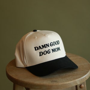 May include: A white and black baseball cap with the text "DAMN GOOD DOG MOM" embroidered in black on the front.
