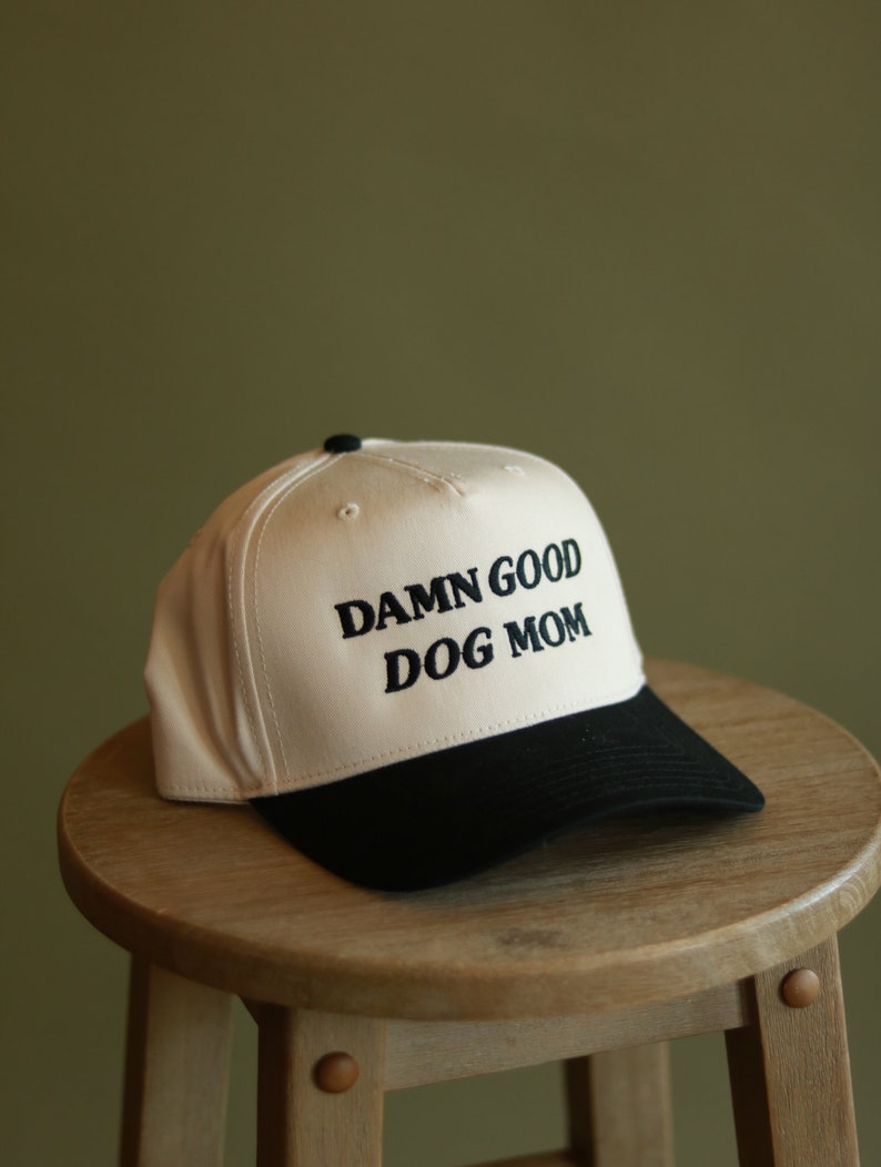 May include: A white and black baseball cap with the text "DAMN GOOD DOG MOM" embroidered in black on the front.
