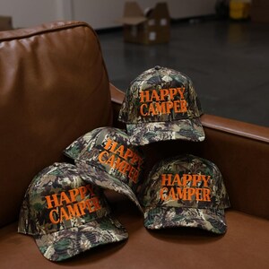 May include: Three baseball caps featuring a camouflage pattern in shades of green, brown, and black. Each cap has the text "HAPPY CAMPER" embroidered in bright orange thread on the front panel. The caps have a structured crown and a curved brim.