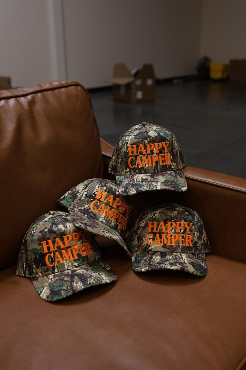 May include: Three baseball caps featuring a camouflage pattern in shades of green, brown, and black. Each cap has the text "HAPPY CAMPER" embroidered in bright orange thread on the front panel. The caps have a structured crown and a curved brim.