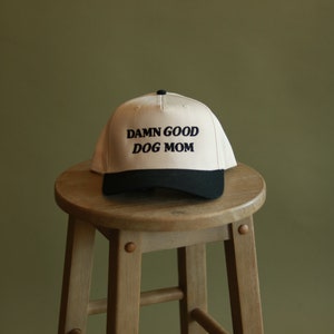 May include: A beige baseball cap with a black brim and the text "DAMN GOOD DOG MOM" embroidered in black.