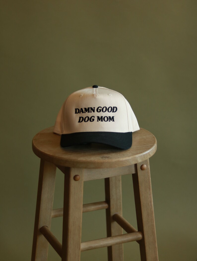 May include: A beige baseball cap with a black brim and the text "DAMN GOOD DOG MOM" embroidered in black.