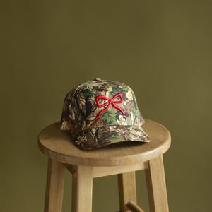 Può includere: Un cappello da baseball mimetico con un fiocco rosso ricamato sul davanti. Il cappello è appoggiato su uno sgabello di legno.