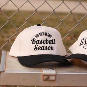 May include: A white baseball cap with a black brim. The front of the cap has black embroidered text that reads "This ain't my first Baseball Season".