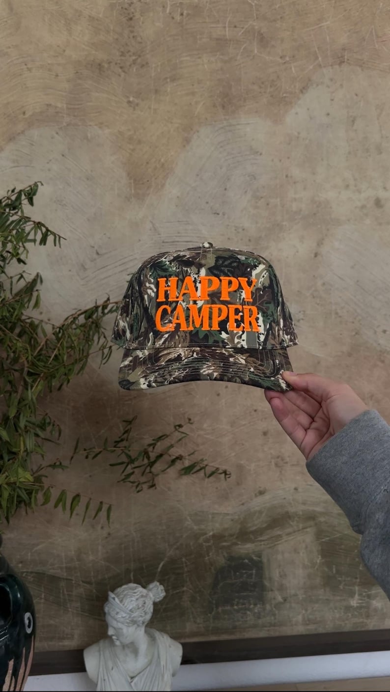 May include: A baseball cap featuring a camouflage pattern in shades of brown, green, and black. The front of the cap displays the text HAPPY CAMPER embroidered in bright orange capital letters. The cap has a structured crown and a curved brim.