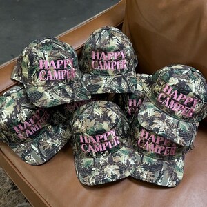 May include: A collection of baseball caps featuring a green, brown, and black camouflage pattern. Each cap has the text "HAPPY CAMPER" embroidered in pink thread on the front panel. The caps are structured with a curved brim and a button top.