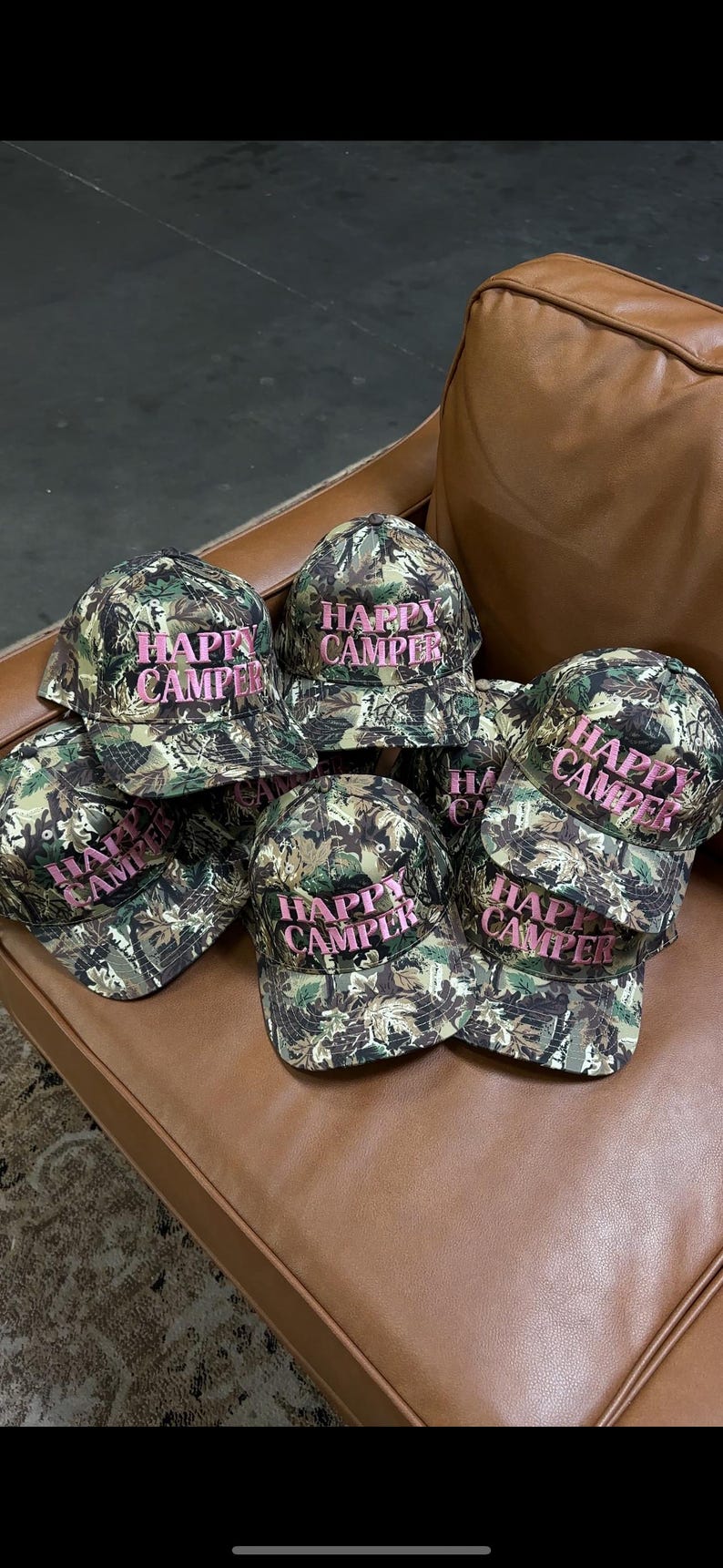 May include: A collection of baseball caps featuring a green, brown, and black camouflage pattern. Each cap has the text "HAPPY CAMPER" embroidered in pink thread on the front panel. The caps are structured with a curved brim and a button top.