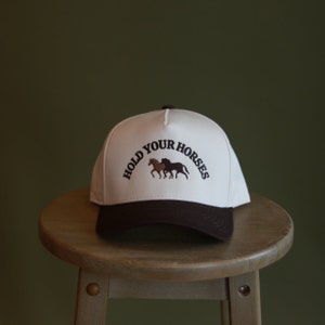 May include: A white and brown baseball cap with the text "HOLD YOUR HORSES" embroidered on the front. The cap features a brown horse silhouette.