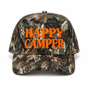 May include: A baseball cap featuring a camouflage pattern in shades of brown, green, and black. The front of the cap displays the text HAPPY CAMPER embroidered in bright orange capital letters. The cap has a structured crown and a curved brim.