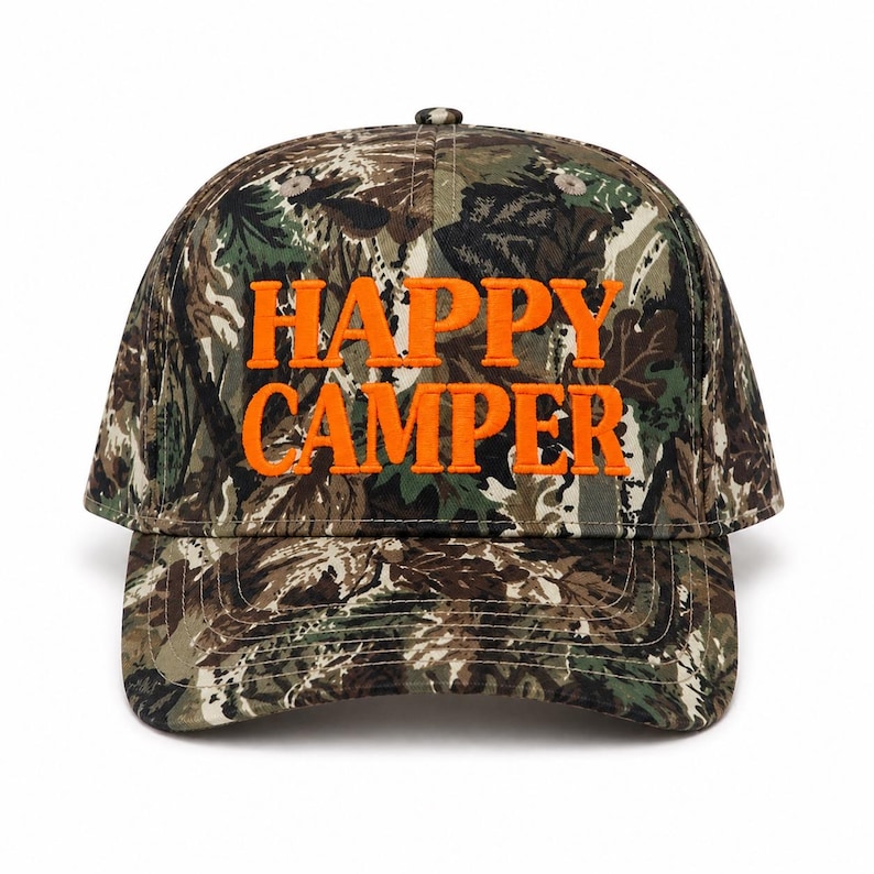 May include: A baseball cap featuring a camouflage pattern in shades of brown, green, and black. The front of the cap displays the text HAPPY CAMPER embroidered in bright orange capital letters. The cap has a structured crown and a curved brim.