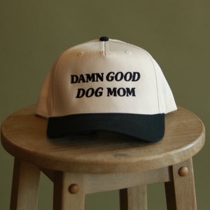 May include: A beige baseball cap with a black brim. The front of the cap has black embroidered text that reads "DAMN GOOD DOG MOM".