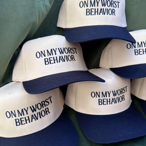 Può includere: Una pila di cappellini da baseball bianchi e blu navy. Ogni cappellino ha un pannello frontale bianco con la frase "ON MY WORST BEHAVIOR" ricamata in blu navy. La visiera e il retro dei cappellini sono blu navy.