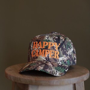 May include: A baseball cap featuring a camouflage pattern in shades of green, brown, and black. The front of the cap displays the text "HAPPY CAMPER" embroidered in bold, bright orange capital letters across the center of the crown.