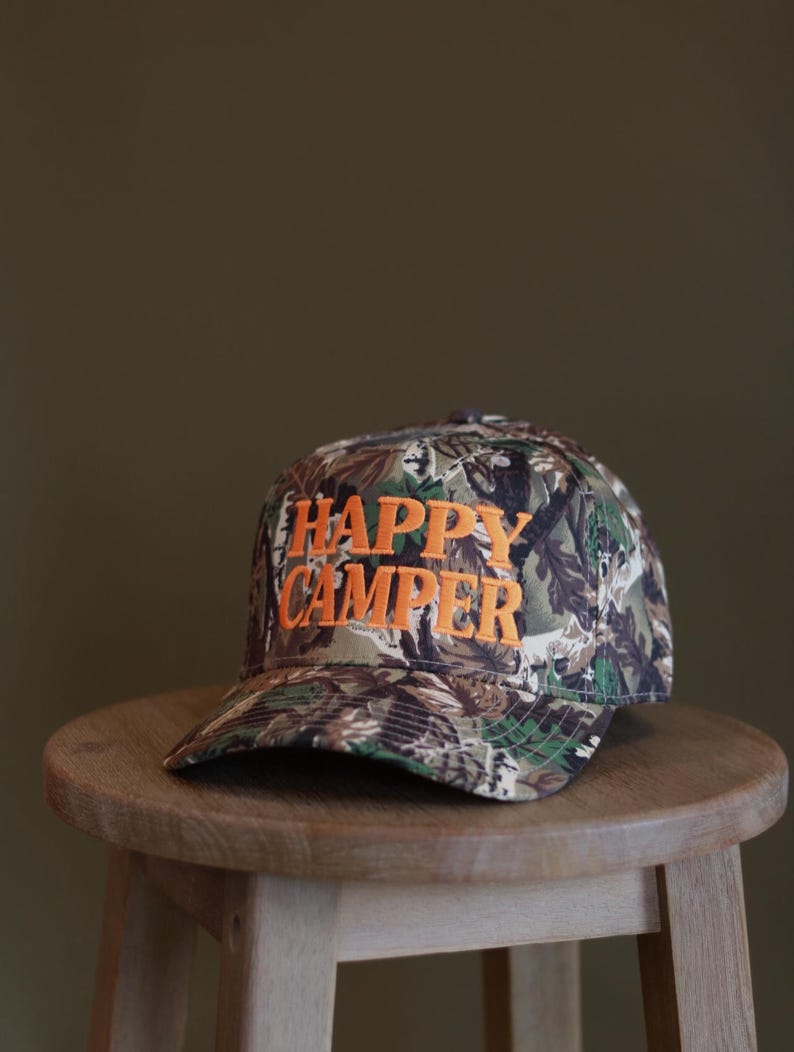 May include: A baseball cap featuring a camouflage pattern in shades of green, brown, and black. The front of the cap displays the text "HAPPY CAMPER" embroidered in bold, bright orange capital letters across the center of the crown.