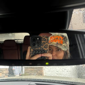 May include: A camouflage baseball cap with the text HAPPY CAMPER embroidered in bright orange thread. The cap features a traditional structured crown and a curved brim, displaying a woodland-style pattern in shades of green, brown, and black.
