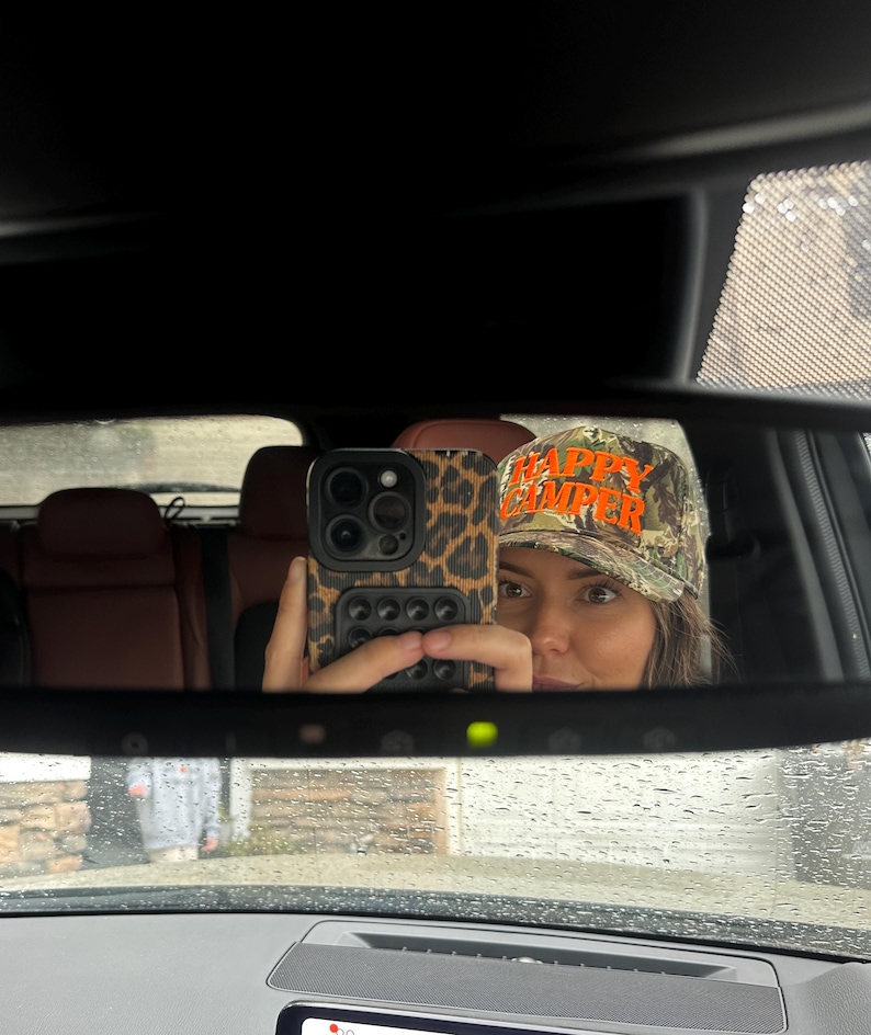 May include: A camouflage baseball cap with the text HAPPY CAMPER embroidered in bright orange thread. The cap features a traditional structured crown and a curved brim, displaying a woodland-style pattern in shades of green, brown, and black.